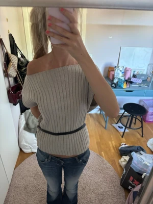 Beige ribbad offshoulder topp - Trendig beige ribbad offshoulder topp med svart bälte i midjan. Toppen har en croppad passform och är perfekt för dig som vill ha en stilren men ändå unik look. Passar snyggt till både jeans och kjol.