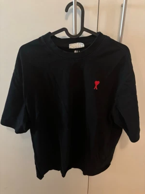 Svart t-shirt - Svart t-shirt från AMI med klassisk passform och korta ärmar. På bröstet finns ett litet rött hjärta med bokstaven A, vilket är varumärkets ikoniska detalj. Tillverkad i mjuk bomull som känns skön mot huden. Lite oversized