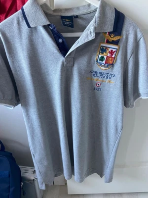 Grå pikétröja från Aeronautica Militare - Snygg grå pikétröja från Aeronautica Militare med broderad logga och emblem på bröstet och höger ärm. Tröjan har korta ärmar, krage med mörkblå kant och knappar framtill. Perfekt för en stilren och sportig look till sommaren eller andra tillfällen. Endast använd 2 gånger. Hör av er vid frågor!