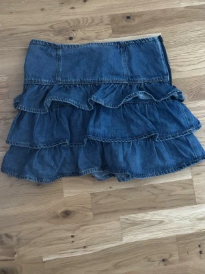 Volangkjol i denim - Säljer en blå jeanskjol med flera volanglager som ger en cool och avslappnad look. Kjolen är tillverkad i denim och har en hög midja. Perfekt för dig som gillar en trendig och ungdomlig stil.