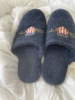 Mörkblå tofflor från Gant - Mysiga mörkblå tofflor från Gant med broderad logga och text på ovansidan. Tofflorna har en mjuk, lurvig insida och slitstark gummisula med mönster. Perfekta för att hålla fötterna varma hemma.