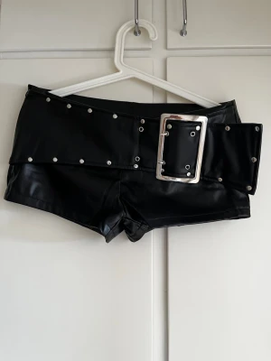 Svarta shorts med bredt bälte - Coola svarta shorts i skinnimitation med bredt bälte och stora silvriga nitar och spänne. Perfekt statement-plagg för dig som vill sticka ut. Snygg passform och trendig look.
