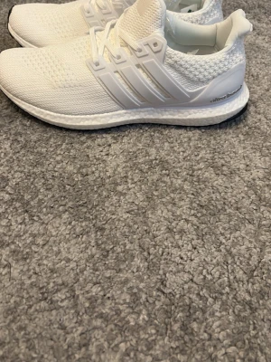 Vita Adidas Ultraboost sneakers - Fräscha helvita Adidas Ultraboost sneakers med stickad ovandel och klassiska tre ränder på sidan. Skorna har en bekväm Boost-sula och ventilerande meshmaterial som ger en sportig och clean look. Perfekta för dig som gillar stilrena och bekväma sneakers.