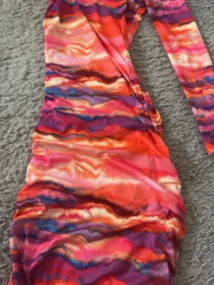 Färgstark långärmad klänning - Säljer en färgstark långärmad klänning med abstrakt mönster i orange, rosa, lila och blå toner. Klänningen har åtsittande passform och rynkade detaljer längs sidorna. Perfekt för dig som vill sticka ut med en unik look.