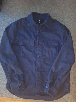 Mörkblå overshirt från H&M - Stilren mörkblå overshirt från H&M med klassisk krage och två stora bröstfickor med lock. Skjortan har knäppning framtill och vid ärmsluten. Tillverkad i ett mjukt, tyg som ger en avslappnad look och bra till alla väder.skriv om du har frågor eller vill se mera bilder ✅