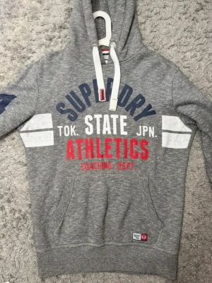 Grå hoodie från Superdry - Säljer en grå hoodie från Superdry med klassisk huva och mjukt, . Hoodien har en enkel design utan tryck på framsidan och är perfekt för en avslappnad stil. Märkeslapp med Superdry Track & Field på insidan.