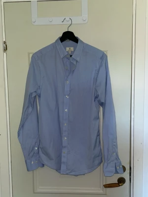 Ljusblå skjorta från Gant - Klassisk ljusblå skjorta från Gant med lång ärm och knappar framtill. Skjortan har en stilren och tidlös design, perfekt till jeans eller chinos. Tillverkad i bomull för en skön känsla och snygg passform.