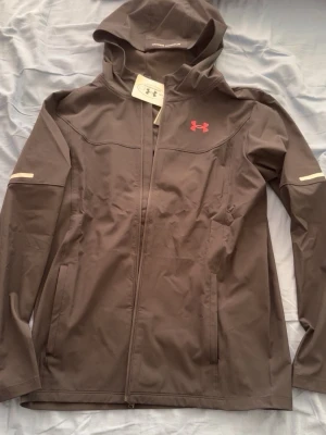 Svart vindjacka från Under Armour - Snygg svart vindjacka från Under Armour med huva och dragkedja framtill. Jackan har diskreta reflexdetaljer på ärmarna och rosa logga på bröstet. Tillverkad i lätt och vattenavvisande material, perfekt för blåsiga dagar.