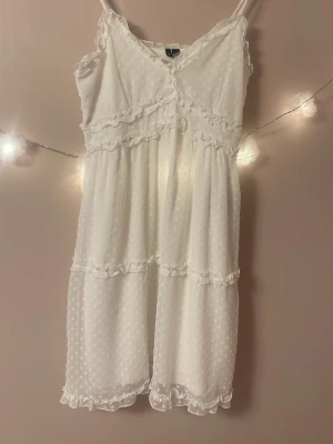 Vit prickig sommarklänning med volanger - Söt vit klänning med tunna axelband och volangdetaljer. Klänningen har ett diskret prickigt mönster och är tillverkad i ett lätt, luftigt material som passar perfekt för varma dagar. Den har en feminin siluett och volanger längs både midja och fåll. Klänningen är från vero Moda och är i storlek L men passar även M. Superfin för skolavslutning,midsommar eller som student klänning💗