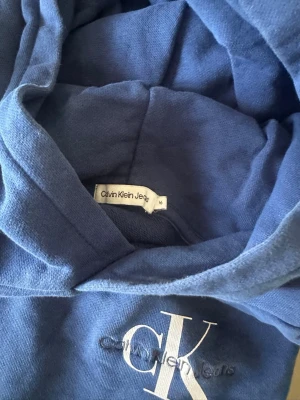 Blå hoodie från Calvin Klein Jeans - Snygg blå hoodie från Calvin Klein Jeans med stor CK-logga broderad på bröstet. Hoodien har en klassisk huva och är tillverkad i mjukt bomullsmaterial. St 170/16 år från kidsbrandstore. 