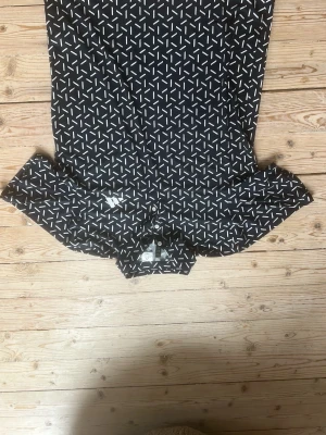 Mönstrad skjorta från Adidas - Snygg mörkblå skjorta från Adidas med vitt grafiskt mönster. Skjortan har klassisk krage, knäppning framtill och långa ärmar. Tillverkad i ett lätt och bekvämt material som passar perfekt för en trendig och sportig look. (Oanvänd).