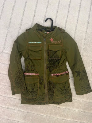 Vintage desigual jacka barn - Stilren grön desigual jacka i storlek 5-6 år med broderade detaljer, paljetter och tryck både fram och bak. Jackan har fyra fickor, dragkedja framtill och dekorativa sömmar i orange. Perfekt skick trotts att den är 10 år gammal, unik design.