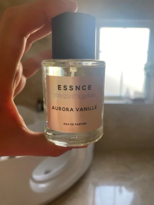 Aurora Vanille parfym från Essnce - Aurora Vanille från Essnce är en Eau de Parfum i en rund, genomskinlig glasflaska med svart lock. Etiketten är ljus med tydlig svart text och parfymen har en stilren och modern design. Doften har en tydlig vaniljkaraktär som känns både söt och mjuk.