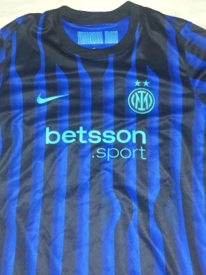 Inter Milan matchtröja blå/svart Nike - Säljer en Inter Milan matchtröja i blått och svart med vertikala ränder. Tröjan har klubbmärke och två stjärnor på bröstet, Nike-logga samt sponsortryck 'betsson sport' framtill. Tillverkad i lätt och ventilerande material, perfekt för fotboll eller träning.