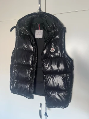 Svart dunväst från Moncler - Snygg svart dunväst från Moncler med glansig finish och klassisk logga på bröstet. Västen har huva, dragkedja framtill och quiltad design som ger en cool och sportig look. Perfekt för lager-på-lager-stil under kyliga dagar. 