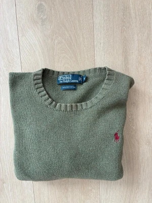 Olivgrön stickad tröja Polo Ralph Lauren - Snygg olivgrön stickad tröja från Polo Ralph Lauren med rund halsringning och ribbade muddar. Tröjan har den klassiska röda logotypen broderad på bröstet och är tillverkad i mjukt material, perfekt för lager på lager och stilrena outfits.