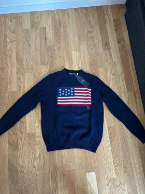 Mörkblå stickad tröja Polo Ralph Lauren - Mörkblå stickad tröja från Polo Ralph Lauren med amerikansk flagga i rött, vitt och blått på bröstet. Rund halsringning och ribbade muddar vid ärmslut och nederkant. Klassisk och stilren design med PL-broderi.