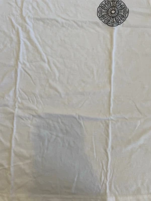 Vit t-shirt med stor svart mandala - Vit t-shirt i bomull med en stor svart mandala-liknande design på ryggen och en mindre likadan detalj på bröstet. Klassisk passform och korta ärmar. Perfekt för dig som gillar grafiska tryck och enkel stil.