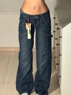 Mörkblå lowwaist baggy jeans med kontrastsömmar - Säljer ett par mörkblå jeans med bred passform och fina detaljer. Jeansen har låg midja, stora fickor fram och bak samt coola detaljer med nitar och dekorativa sömmar på bakfickorna. Lappen finns kvar på