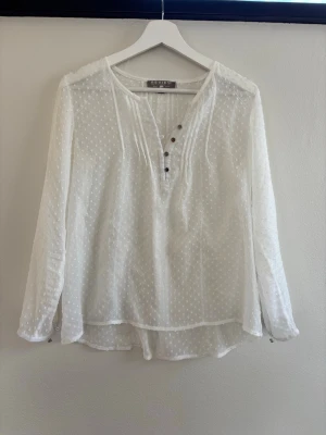 Vit prickig blus från Review - Söt vit blus från Review med diskret prickigt mönster och tunnt, lätt material. Blusen har lång ärm, rundad halsringning och knappar framtill i olika färger. Perfekt för en avslappnad men ändå stilren look.