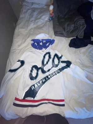 Vit hoodie från Polo Ralph Lauren - Säljer en vit hoodie från Polo Ralph Lauren med stor logga i mörkblått och silver över bröstet. Huvan har blått innerfoder med vita stjärnor och nederkanten har röda och mörkblå ränder. Perfekt för dig som gillar streetwear och vill sticka ut.