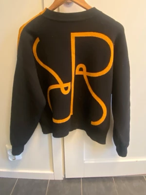 Rodebjer Tröja - Stickad tröja "Reilly" från Rodebjer. Rundad hals och ballongärm. Storlek & passform: S men går till M (jag har M)  Material: 65% Viskos, 28% Polyester, 7% Polyamid.Cool svart stickad tröja med ett stort grafiskt orange mönster på ryggen. Tröjan har rund halsringning och långa ärmar. Perfekt för dig som gillar statement-plagg och vill sticka ut lite extra.