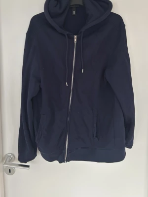 Mörkblå hoodie med dragkedja - Säljer en mörkblå hoodie med dragkedja framtill och två fickor. Hoodien har huva med dragsko och ribbade muddar vid ärmslut och nederkant. Perfekt för en avslappnad och stilren look.