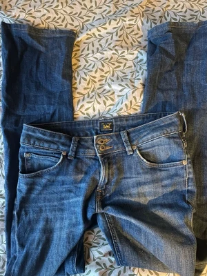 Blå jeans från Lee med dubbelknapp - Säljer ett par klassiska blå jeans från Lee med snygg dubbelknapp framtill och femficksdesign. Jeansen har normal passform och är gjorda i bomull. Perfekt för dig som gillar tidlös denimstil och vill ha ett par jeans som funkar till allt. W27 L31