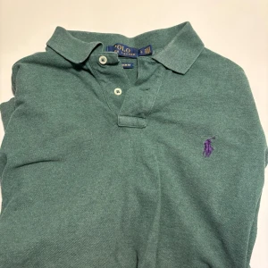Grön pikétröja från Polo Ralph Lauren - Klassisk grön pikétröja från Polo Ralph Lauren med lila broderad logga på bröstet. Tröjan har korta ärmar, krage och knappar vid halsen. Perfekt till en avslappnad men ändå stilren look.