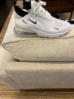 Nike Air Max 270 vita sneakers - Nike Air Max 270 sneakers i vitt med svarta detaljer och synlig luftbubbla i hälen. Ovandelen är i mesh för bra ventilation och skon har en sportig, modern look. Perfekt för dig som gillar streetwear och vill ha bekväma skor med snygg design.