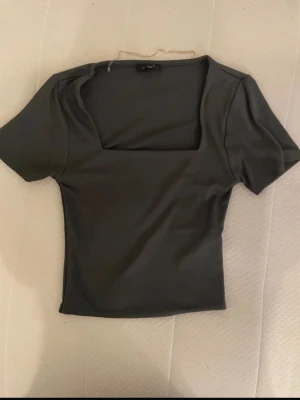 Mörkgrå topp med fyrkantig ringning - Snygg mörkgrå topp med korta ärmar och fyrkantig ringning. Modellen är figurnära och croppad, perfekt till höga jeans eller kjol. Enkel och stilren design som passar till många olika looks.