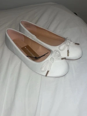 Vita ballerinaskor från Anna Field - Snygga vita ballerinaskor från Anna Field med rund tå och dekorativ rosett framtill. Skorna har en klassisk siluett och är tillverkade i återvunnen polyester med mjuk innersula i beige. Perfekta för en stilren och enkel look. Prislapp kvar!! 