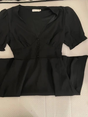 Svart klänning med puffärm och knappar - Snygg svart klänning med puffärmar och v-ringning. Klänningen har dekorativa knappar framtill och en markerad midja som ger en feminin siluett. Perfekt för dig som gillar klassisk stil med moderna detaljer.