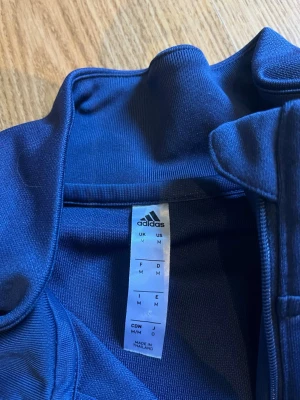 Blå Sverige träningströja Adidas M - Säljer en blå långärmad träningströja från Adidas med Sveriges landslagsemblem på bröstet. Tröjan har vita ränder på ärmarna, halv dragkedja och loggor från ICA och Bauhaus. Tillverkad i funktionsmaterial, perfekt för träning eller uppvärmning.