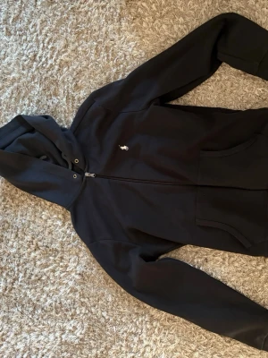 Svart hoodie från Polo Ralph Lauren - Snygg svart hoodie med dragkedja från Polo Ralph Lauren. Klassisk design med huva, fickor framtill och den ikoniska lilla loggan broderad på bröstet. Perfekt för en chill och stilren look. Tillverkad i mjukt bomullsmaterial.