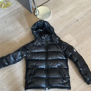 Moncler Maya puffer - Säljer en svart glansig dunjacka från Moncler med huva och dragkedja. Jackan har två framfickor med dragkedja och en ärmficka med Moncler-logga. Klassisk pufferstil som håller värmen under kalla dagar. En defekt. 