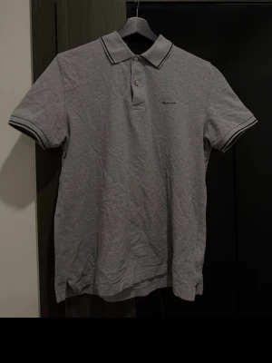 Grå pikétröja från GANT - Grå pikétopp från GANT med korta ärmar, klassisk krage och två knappar framtill. Broderad logga på bröstet och mörkblå detaljer på krage och ärmslut. Tillverkad i mjuk bomull.