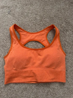 Orange sport-bh från NVGTN - Snygg orange sport-bh från NVGTN med brottarrygg och ribbad nederkant. Tillverkad i stretchigt material som ger bra stöd under träningen. Logotypen är tryckt i vitt på ryggen. Perfekt för gymmet eller löprundan. Använd endast en gång💗