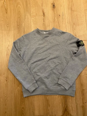 Grå sweatshirt från Stone Island - Klassisk grå sweatshirt från Stone Island med rund halsringning och ribbade muddar. Tröjan har lång ärm och den ikoniska Stone Island-loggan på vänster ärm. Perfekt för en avslappnad och stilren look.