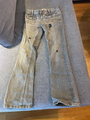 Grå slitna bootcut jeans - Säljer ett par grå bootcut jeans med slitningar och hål för en edgy look. Jeansen har klassiska bakfickor och är tillverkade i denim. Egna klippta hål.(pris kan diskuteras☺️)