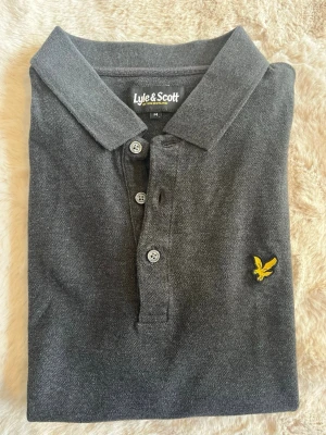 Pikétröja från Lyle & Scott - Klassisk mörkgrå pikétröja från Lyle & Scott med korta ärmar och krage. Tröjan har tre knappar framtill och den ikoniska gula fågel-loggan broderad på bröstet. 