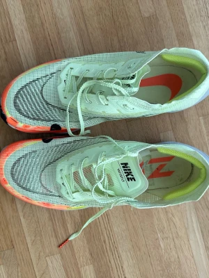 Nike Vaporfly ljusgröna sneakers - Nike Vaporfly sneakers i ljusgrönt meshmaterial med inslag av orange och svart på sulan. Skorna har en sportig design, synlig logga på plösen och innersulan, samt en lätt och ventilerande ovandel. Sulan är tjock och designad för löpning med extra dämpning.