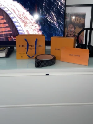 Svart rutigt Louis bälte - Snyggt svart bälte från Vuitton med klassiskt rutigt mönster och ikoniskt V spänne. Bältet har en stilren design som passar till jeans och byxor. Tillverkat i skinn med en matt finish och diskreta detaljer. Storlek 110cm