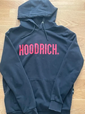 Svart HOODRICH hoodie med röd logga - Svart hoodie från HOODRICH med stor röd logga på bröstet. Klassisk modell med huva och dragsnören samt magficka framtill. Tillverkad i mjukt sweatshirtmaterial som är perfekt för en avslappnad streetwear-look.