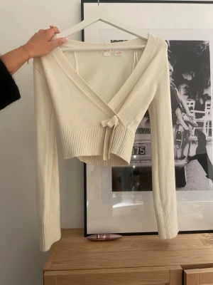 Krämvit omlottstickad tröja - Supersnygg krämvit omlottstickad tröja med långa ärmar och bred ribbad kant i midjan. Perfekt att styla med jeans eller kjol för en trendig look. Storleken är 152cm men är snygg att ha kort med ett vitt linne