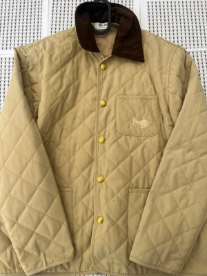 Beige quiltad cardiganjacka med bruna detaljer - Quiltad beige cardiganjacka från Celine med bruna manchesterdetaljer på kragen och guldfärgade knappar. Jackan har en bröstficka med broderad fisk, två sidofickor och ett klassiskt rutmönster. Perfekt för dig som gillar stilren och avslappnad look.