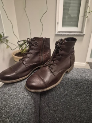 Rosa Negra Bruna läderstövletter med snörning 37 - Snygga bruna stövletter i äkta läder med snörning framtill och dragkedja på sidan. Modellen har rund tå och en låg klack, perfekt för dig som gillar klassisk och stilren design. Yttersulan är ljus och ger en cool kontrast mot det mörka lädret.