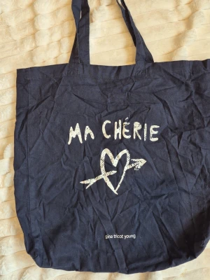 Blå tygväska Ma Chérie Gina Tricot - Cool marinblå tygväska från Gina Tricot Young med trycket 'MA CHÉRIE' och ett hjärta med pil på framsidan. Perfekt för att bära böcker, laptop eller shopping. Långa axelband gör den enkel att slänga över axeln. Materialet är bomull.