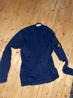 Mörkblå långärmad tröja Stone Island - Mörkblå långärmad tröja från Stone Island med klassisk rund halsringning och ikonisk logotyp-patch på vänster ärm. Tröjan är i mjukt material och har en stilren, enkel design som passar perfekt till jeans eller chinos.