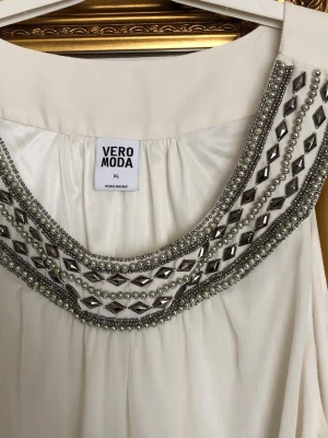 Vit ärmlös topp med pärldetaljer Vero Moda - Säljer en vit ärmlös topp från Vero Moda i storlek XL. Toppen har ett brett smockband nedtill och är dekorerad med silverfärgade pärlor och stenar runt den runda halsringningen. Perfekt för dig som gillar detaljer och en luftig passform.  Längd: 73cm och armhåla till armhåla 60cm. Nytt aldrig använt. 75kr
